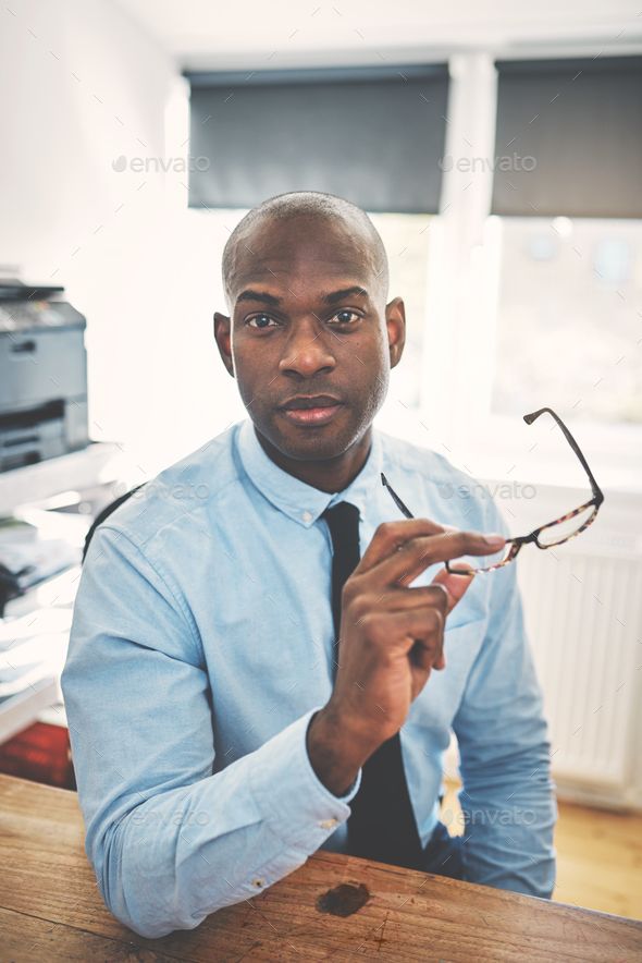 Confident African businessman in a shirt and tie holding glasses while sitting at a desk in his home office